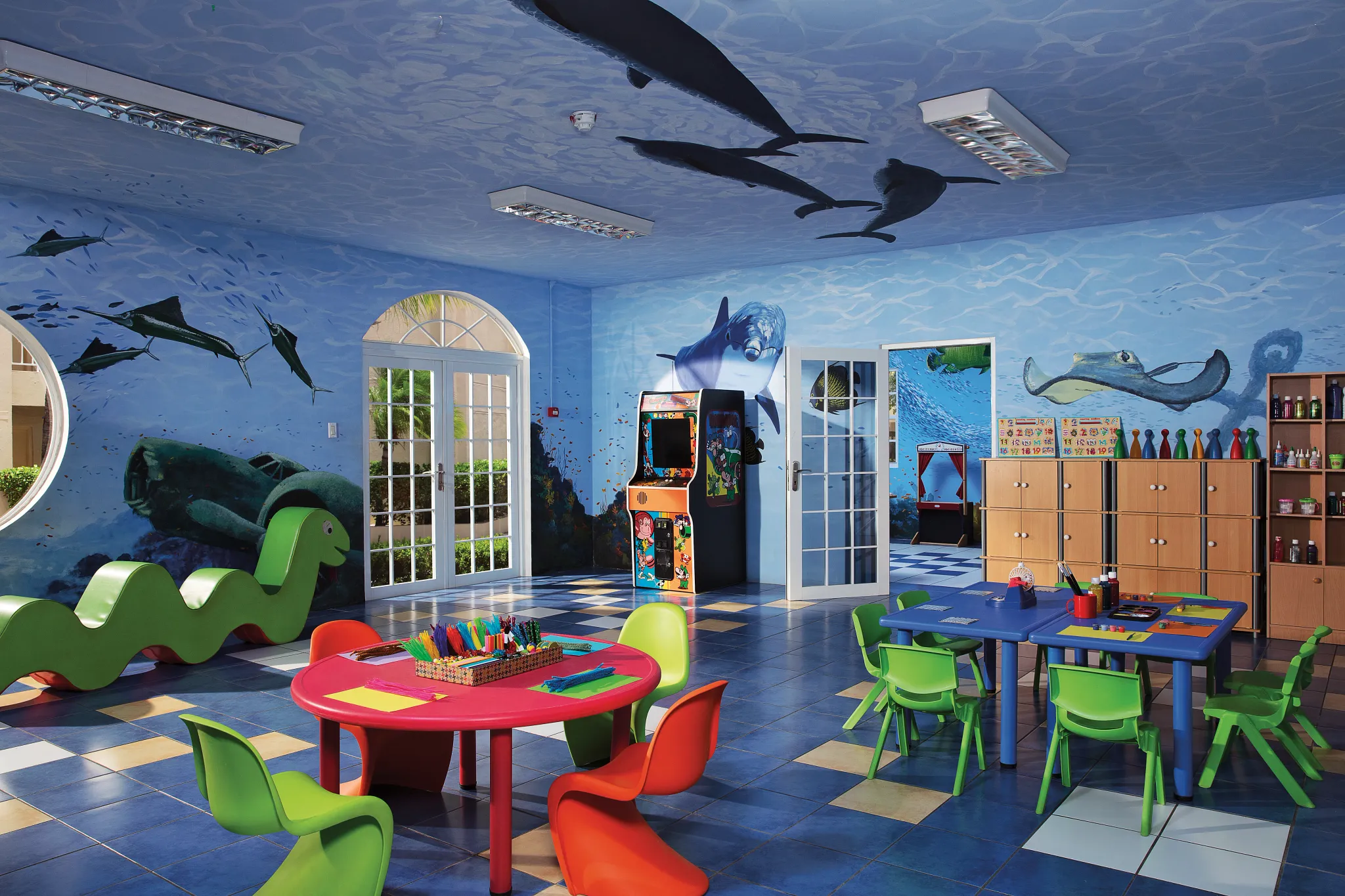 Colorful indoor kids playroom with ocean-themed murals, craft tables, and arcade games at Sunscape Curaçao Resort, Spa & Casino.